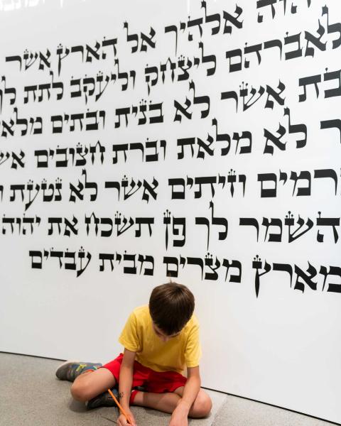 Child sits in front of a wall with Hebrew writing and writes on a piece of paper.